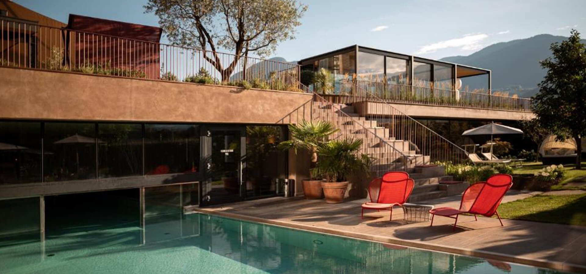 Modernes Haus mit großer Glasveranda und Terrasse, Außenpool im Vordergrund, zwei rote Liegestühle am Beckenrand, Treppe zur oberen Gartenterrasse und Bergkulisse.