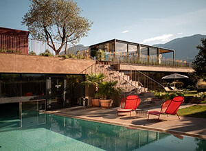 Modernes Haus mit großzügiger Terrasse und klarem Swimmingpool im Vordergrund; zwei rote Gartenstühle neben dem Pool, Pflanzen und Sonnenschirm, obere verglaste Wohnetage mit Treppe, Berge im Hintergrund.