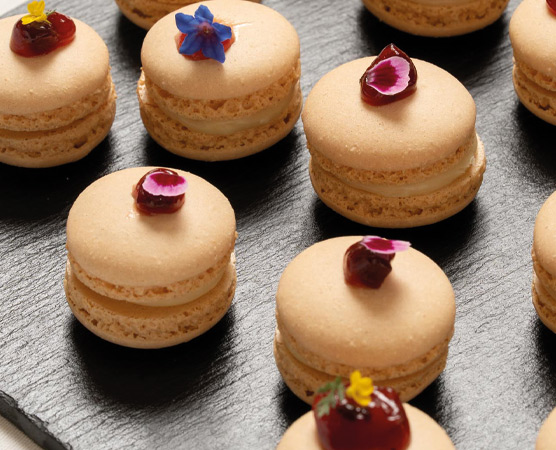 Auf einem schwarzen Servierbrett liegen mehrere Macarons. Diese bestehen aus goldbrauner Baiser-Hülle und sind mit einer Cremefüllung gefüllt. Einige Macarons sind mit Essblumen und kleinen Fruchtstücken dekoriert, die ihnen eine farbenfrohe Note verleihen.