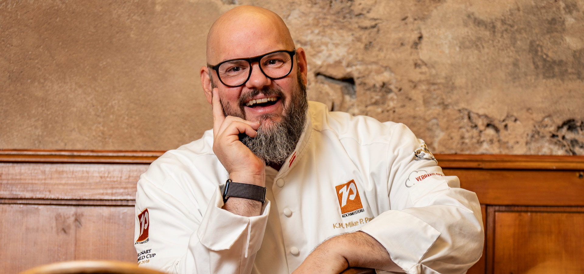 Ein lächelnder Koch in einer weißen Kochjacke sitzt entspannt. Er trägt eine Brille und hat einen dichten Bart. Der Hintergrund zeigt eine rustikale Wand aus Stein, die eine gemütliche Atmosphäre schafft.