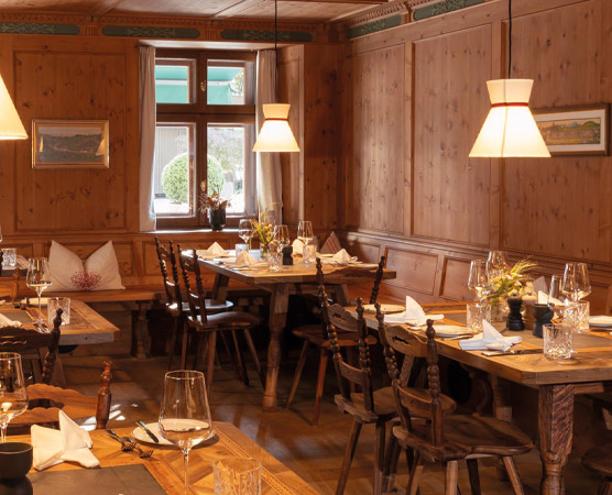 Ein gemütlicher, holzverkleideter Raum mit mehreren Tischen und Stühlen. Auf den Tischen sind Gläser, Besteck und Servietten sorgfältig gedeckt. An der Wand hängen Bilder, und sanfte Beleuchtung schafft eine einladende Atmosphäre. Ein Fenster bietet Blick nach draußen.