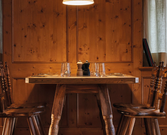Ein holzvertäfelter Raum mit einem rustikalen Tisch in der Mitte. Auf dem Tisch stehen zwei Gläser, ein Salz- und Pfefferstreuer und zwei Gedecke. Rund um den Tisch stehen zwei Holzstühle. Eine Hängeleuchte sorgt für sanfte Beleuchtung.