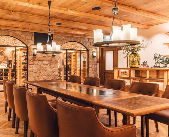 Ein eleganter Speise- oder Besprechungsraum mit einem langen Holztisch und bequemen Stühlen. Über dem Tisch hängen moderne Lichtquellen. Im Hintergrund sind Weinkeller mit verschiedenen Flaschen und eine dekorative Ecke sichtbar, die eine einladende Atmosphäre schafft.