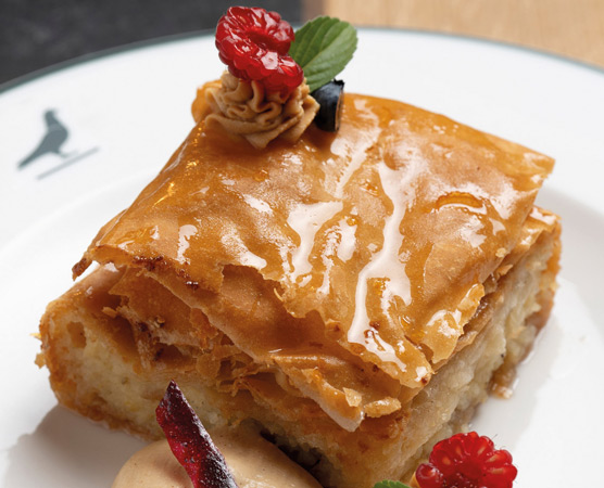 Ein Stück Baklava auf einem Teller, mit goldener, knuspriger Oberseite. Es ist garnisiert mit einer kleinen Rosette aus Sahne, einem Blatt Minze und roten Beeren. Daneben liegt eine Portion Eiscreme.