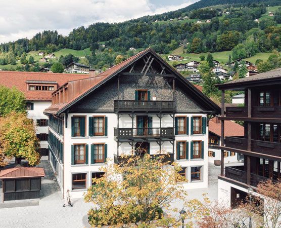 Ein traditionelles Gebäude mit weißer Fassade und dunklem Dach steht im Vordergrund. Umgeben von Bäumen und angrenzenden modernen Gebäuden, zeigt die Szene die malerische Architektur in einer hügeligen Landschaft. Im Hintergrund sind sanfte Hügel und weitere Wohngebäude sichtbar.