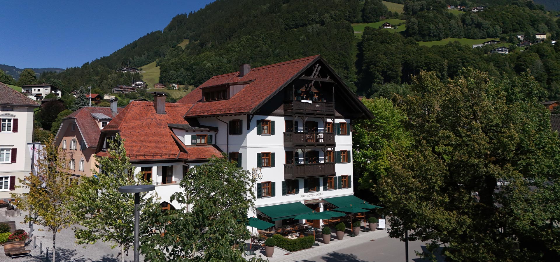 Ein traditionelles Hotel mit einem hellen, weiß gestrichenen Gebäude und roten Ziegeldächern. Es ist von Bäumen und einer schönen Landschaft umgeben, die Hügel und Wälder umfasst. Auf der Terrasse des Hotels stehen grüne Sonnenschirme und Tische für Gäste.