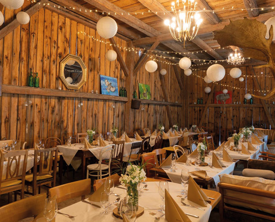 Ein rustikales Restaurant mit Holzwänden und einem warmen Ambiente. Tische sind elegant gedeckt mit Blumenarrangements und Stoffservietten. Lichterketten und Laternen sorgen für eine gemütliche Beleuchtung. Gemälde und Dekorationen an den Wänden runden die Atmosphäre ab.