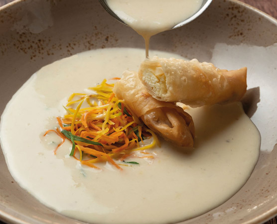 Ein Teller mit einer cremigen Suppe, auf dem eine geschmackvolle Füllung ist, die mit knusprigem Teig umhüllt ist. Die Suppe ist mit buntem, feingeschnittenem Gemüse garniert, das aus gelben und orangefarbenen Streifen besteht.