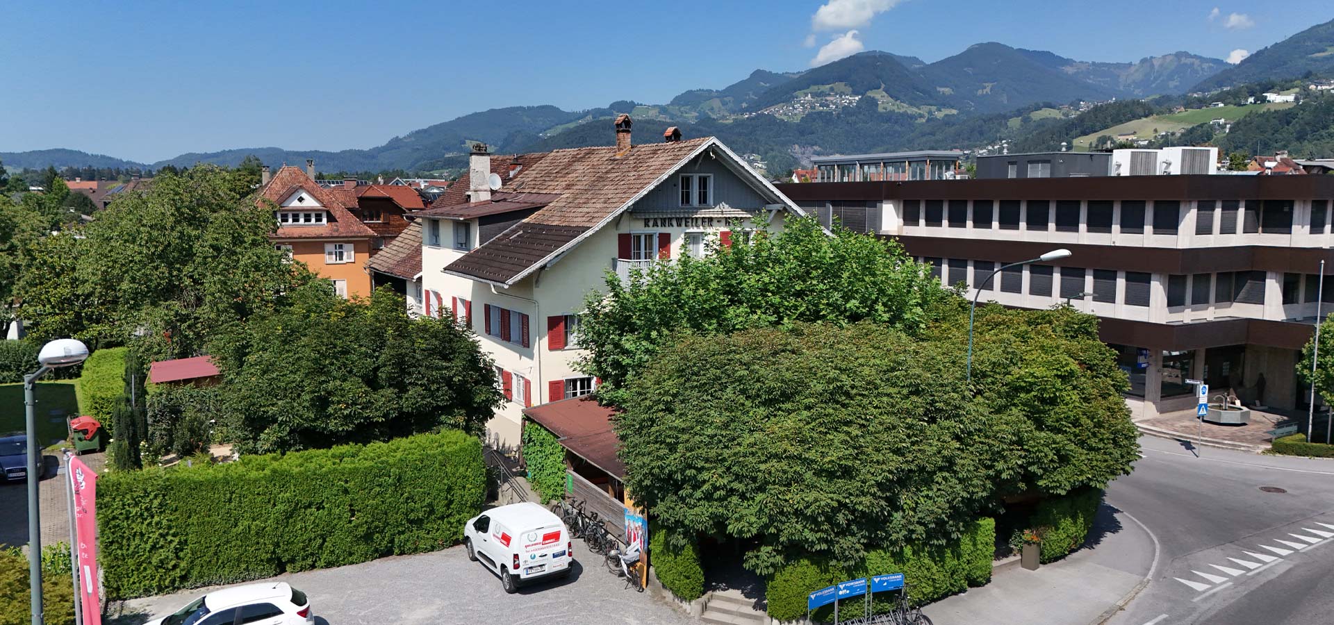 Das Bild zeigt eine städtische Umgebung mit verschiedenen Gebäuden. Ein helles zweigeschossiges Haus mit roten Fenstern und einem angrenzenden Garten ist prominent. Im Hintergrund erheben sich grüne Berge unter einem klaren blauen Himmel. Ein Fahrzeug steht auf einem gepflasterten Parkplatz.