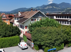Ein mehrstöckiges Haus mit rotem Fensterrahmen, umgeben von hohen Bäumen und buschigen Hecken. Im Vordergrund steht ein weißes Fahrzeug. Im Hintergrund sind grüne Hügel und Berge zu sehen, die den ländlichen Charakter der Umgebung unterstreichen.