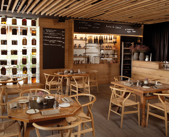 Ein gemütliches Restaurant mit Holzmöbeln und Tischen. An der Wand sind flascheweise verschiedene Spezialitäten ausgestellt. An der Theke stehen Getränke und hübsche Blumenarrangements. Eine einladende Atmosphäre betont den Fokus auf Genuss und geselliges Beisammensein.