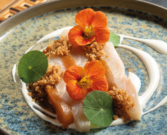 Ein kunstvoll angerichteter Teller mit zarten Fischscheiben, garniert mit orangefarbenen Blüten und grünen Blättern. Der Boden des Tellers ist mit einer knusprigen Krümel-Textur dekoriert, und ein feiner, cremiger Belag umrahmt das Gericht.