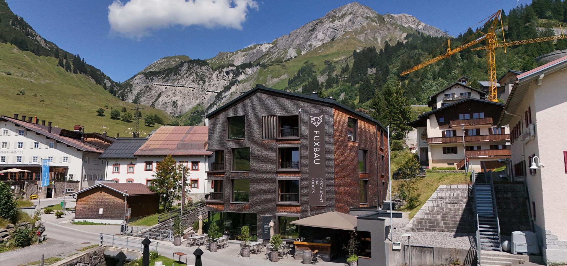 Ein modernes Hotelgebäude mit dem Namen „Fuxbau“ steht im Vordergrund, umgeben von Bergen und grünen Wiesen. Das Hotel hat große Fenster und eine Terrasse mit Sitzmöglichkeiten. Im Hintergrund sind Häuser und ein Kranausleger sichtbar, die die alpine Umgebung betonen.