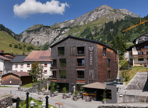 Das Bild zeigt das moderne Gebäude des Hotels Fuxbau, umgeben von malerischen Bergen und einer klaren blauen Himmel. Die Architektur kombiniert Holz und Stein, und im Vordergrund sind Außensitzbereiche mit Tischen und Stühlen. Das Hotel liegt in einer ruhigen, alpinen Umgebung.