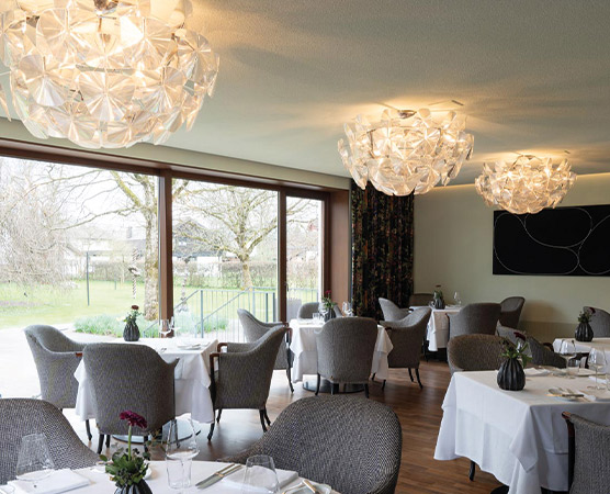 Ein stilvolles Restaurant mit eleganten Tischen und Stühlen. Kreative Lampen hängen von der Decke. Große Fenster bieten einen Blick auf den Außenbereich mit Bäumen. Die Tische sind sauber gedeckt und mit Blumenarrangements dekoriert. Eine angenehme und einladende Atmosphäre ist spürbar.