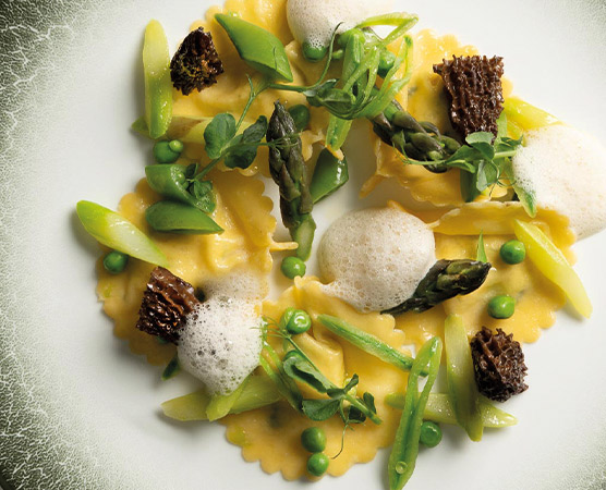 Ein Teller mit Ravioli, garniert mit Spargel, Erbsen und Morchelpilzen. Darüber liegt ein feiner Schaum. Frische Kräuter und einige grüne Gemüsestücke ergänzen das Gericht. Die Anordnung der Zutaten ist kreativ und ansprechend.