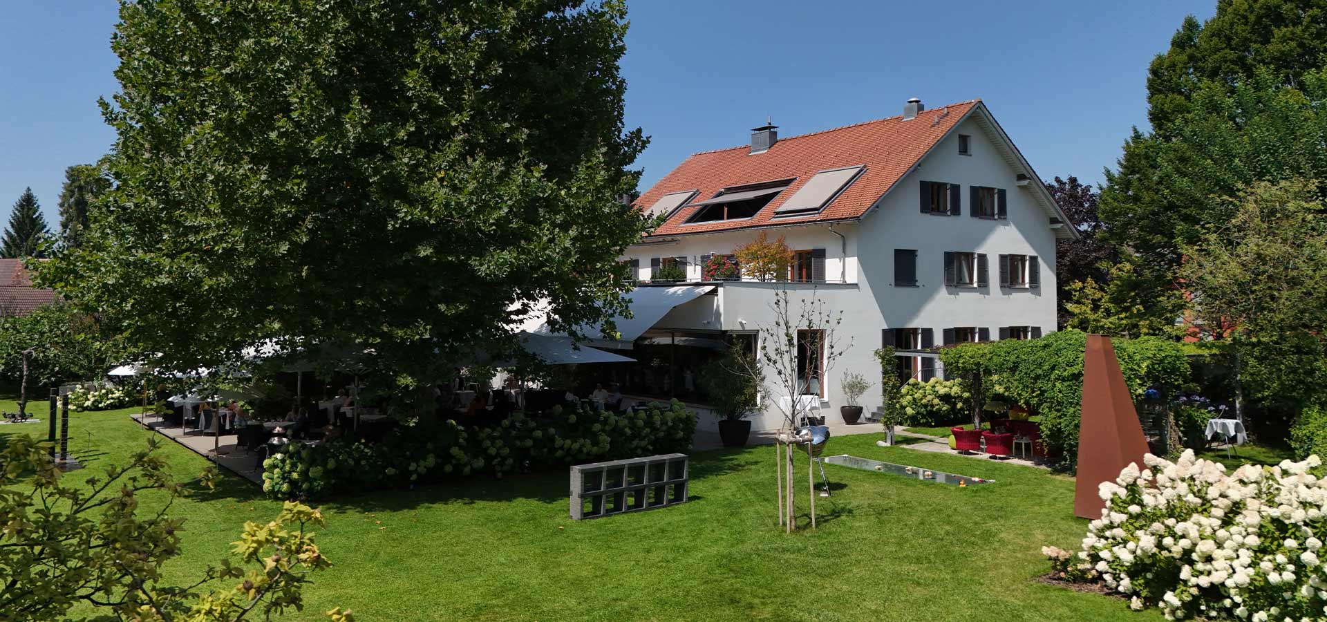 Ein stattliches, helles Gebäude mit einem roten Dach und weißen Wänden steht in einem gepflegten Garten. Im Freien sind Tische und Stühle auf einer Terrasse platziert, umgeben von grünen Pflanzen und Blumen. Sonnenlicht beleuchtet die Szene an einem klaren, blauen Himmel.