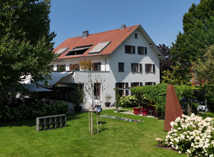 Eine weiße Villa mit roten Dachziegeln in einem gepflegten Garten. Umgeben von Bäumen und Blumen sind verschiedene Sitzgelegenheiten und ein moderner Skulptur im Garten sichtbar. Die Szene vermittelt eine einladende und ruhige Atmosphäre.