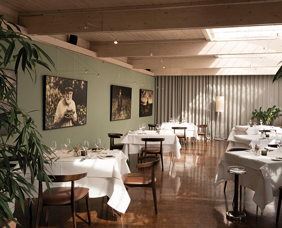 Ein elegantes Restaurant mit hellen Holzwänden und großen Fenstern. Tische sind mit weißen Tischdecken gedeckt, und stilvolle Stühle umgeben die Tische. An den Wänden hängen Kunstwerke, und Pflanzen fügen eine natürliche Note hinzu. Die Atmosphäre ist einladend und geschmackvoll.