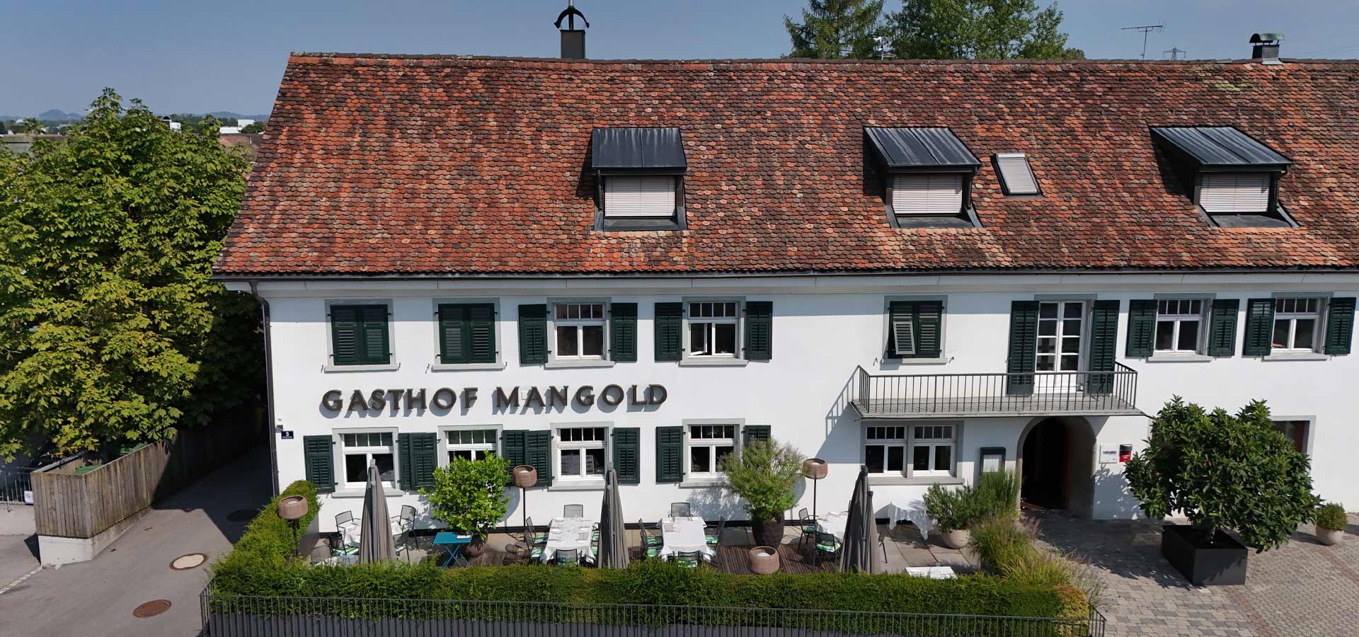 Ein traditionelles Gasthaus mit weißer Fassade und einem roten Ziegeldach. Der Name "Gasthof Mangold" ist sichtbar angebracht. Vor dem Gebäude befinden sich Gartentische, Stühle und Sonnenschirme, umgeben von grünen Pflanzen.