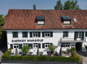 Das Bild zeigt den Gasthof Mangold, ein mehrstöckiges Gebäude mit einem schrägen, roten Ziegeldach. An der Fassade sind grüne Fensterläden und eine Beschilderung mit dem Namen des Gasthofs zu sehen. Vor dem Gebäude befinden sich Terrassen mit Sitzgelegenheiten.