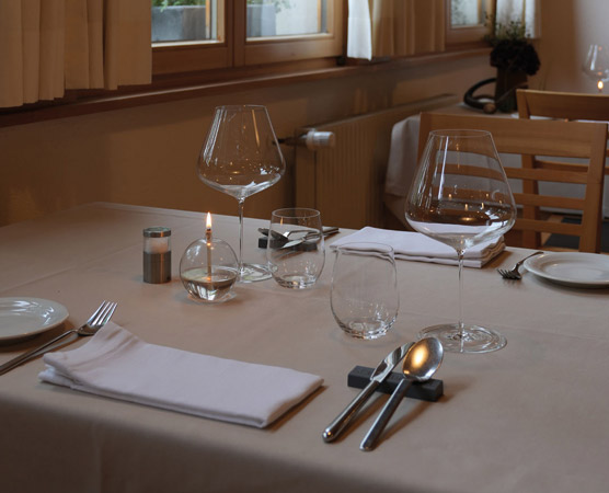 Ein gedeckter Tisch in einem eleganten Restaurant. Auf dem Tisch befinden sich zwei große Weingläser, ein kleiner Kerzenhalter, mehrere Wassergläser, ein Besteckset und gefaltete Servietten. Die Tischdecke ist hell und der Raum wirkt freundlich und einladend.