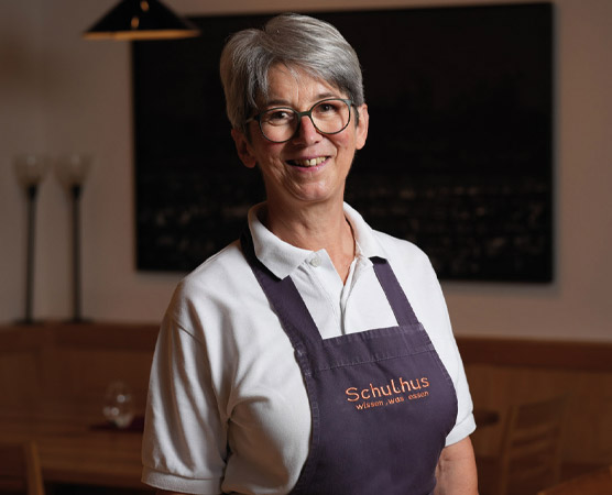 Eine lächelnde Frau mit grauen Haaren und Brille trägt ein weißes Shirt und eine lila Schürze mit dem Aufdruck „Schulhus“. Im Hintergrund sind Tisch und Dekoration eines Restaurants zu sehen. Die Atmosphäre wirkt freundlich und einladend.
