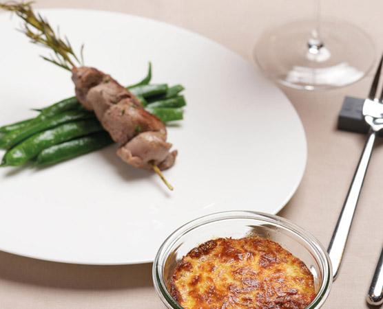 Ein elegantes Gericht auf einem weißen Teller: Spieße mit Fleisch und frischen grünen Bohnen. Daneben ein kleines Glas mit einem herzhaften Auflauf, der goldbraun gebacken ist. Ein Weinglas und Besteck sind im Hintergrund sichtbar.