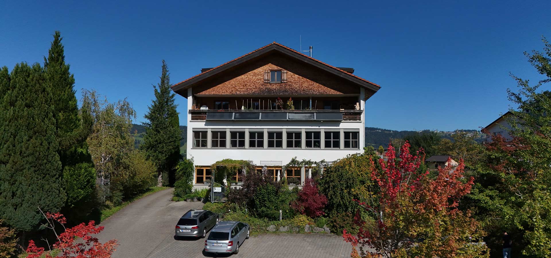 Ein mehrstöckiges Haus mit einem Satteldach, umgeben von Bäumen und buntem Laub. Im Vordergrund sind zwei parkende Autos auf einem gepflasterten Weg zu sehen. Der Himmel ist klar und blau, was auf einen sonnigen Tag hinweist.