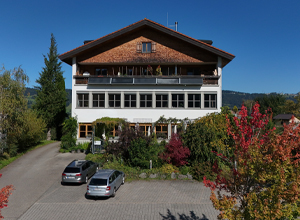 Ein mehrstöckiges Gebäude mit Holzverkleidung und Balkon ist im Vordergrund zu sehen. Um das Haus herum sind einige Bäume und bunte Sträucher sowie ein Parkplatz mit mehreren Autos. Der Himmel ist klar und blau.