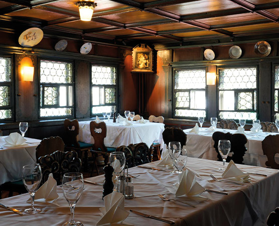 Ein elegantes Restaurant mit Holzdekor, hellen Fenstern und gedeckten Tischen. Die Tische sind mit weißen Tischdecken, Gläsern und gefalteten Servietten dekoriert. An den Wänden sind kunstvolle Teller angebracht, die eine gemütliche, einladende Atmosphäre schaffen.