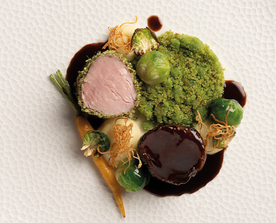 Ein kreativ angerichteter Teller mit zartem Fleisch, umgeben von grünem Püree, Rosenkohl und kleinen Karotten. Eine dunkle Soße verleiht dem Gericht zusätzlichen Kontrast. Die Komposition zeigt verschiedene Texturen und Farben, die eine ansprechende Mahlzeit präsentieren.