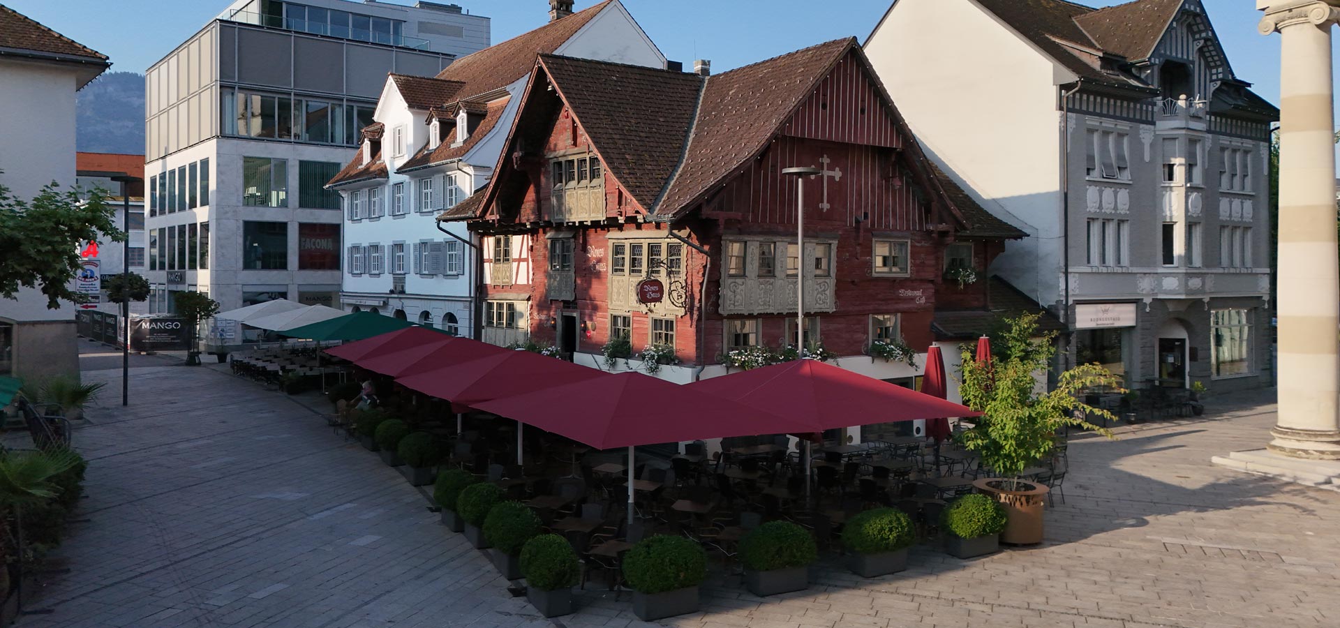 Ein historisches Gebäude mit roten Holzverkleidungen und Fensterläden befindet sich zwischen modernen Bauten. Vor dem Haus gibt es einen Außenbereich mit Tischen und roten Sonnenschirmen. Der Platz ist tagsüber sonnig und einladend gestaltet.