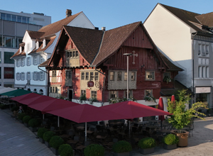 Ein traditionelles, historisches Gebäude in rot und beige mit mittelalterlicher Architektur. Darunter befinden sich Tische und Stühle unter roten Sonnenschirmen im Freien. Im Hintergrund sind moderne Gebäude sichtbar, die einen Kontrast zur altmodischen Bauweise bieten.