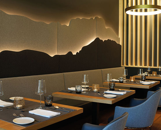Ein modernes Restaurant mit eleganten Holztischen und gepolsterten Sitzbänken. Die Wände sind mit einer stilisierten Berglandschaft dekoriert und sanft beleuchtet. Tischgedecke sind vorbereitet mit Geschirr, Gläsern und Kerzenhaltern, die eine gemütliche Atmosphäre schaffen.