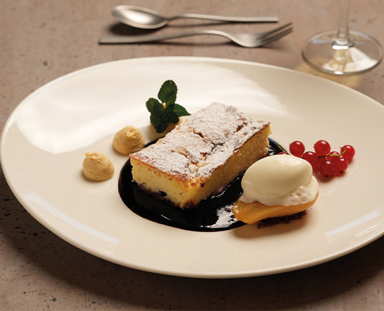 Ein Stück Kuchen mit Puderzucker, serviert auf einem weißen Teller. Daneben befinden sich kleine Portionen von Sahne und einer Fruchtsauce. Rote Johannisbeeren und minzige Blätter garnieren das Dessert, das in einer dunklen Sauce liegt.