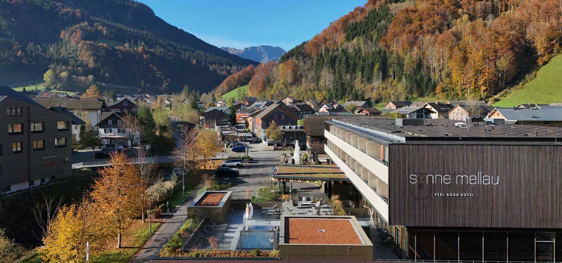 Das Bild zeigt die Umgebung des "Sonne Melau"-Hotels in den Bergen, umgeben von buntem Herbstlaub. Im Vordergrund sind moderne Gebäude und ein Außenbereich mit Sitzgelegenheiten und einem kleinen Pool zu sehen. Die Kulisse wird von sanften Hügeln und bewaldeten Bergen geprägt.