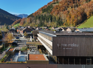 Luftaufnahme des Hotels "Sonne Mellau" in einer bergigen Umgebung. Das moderne Gebäude steht in einem ruhigen Dorf, umgeben von bunten Herbstbäumen und grünen Hügeln. Ein Außenbereich mit Pools ist sichtbar. Die Szenerie vermittelt eine ruhige, einladende Atmosphäre.