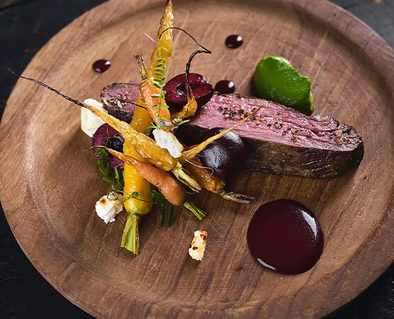 Auf einem runden Holzbrett: eine rosa gebratene Scheibe Rindfleisch, glasierte bunte Babykarotten, grüne Kräuter, kleine Käsebrösel und dunkle Sauce-Tropfen dekorativ angerichtet.