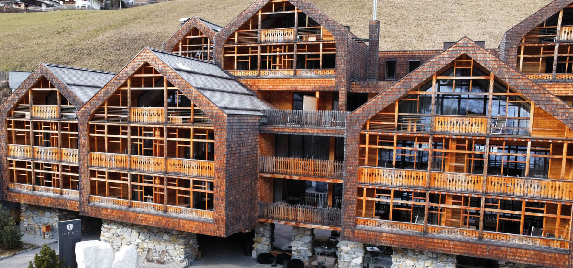 Mehrteilige hölzerne Bergchalet-Anlage mit mehreren Giebeldächern, durchgehenden Holzbalkonen, großen Fenstern und steinernem Sockel vor einem grasigen Hang.