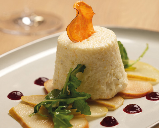 Ein Dessert auf einem weißen Teller: Eine luftige Mousse in Form eines Turms, garniert mit einem knusprigen Gemüsechip. Daneben liegen dünne Scheiben von Äpfeln und ein paar Tropfen einer Fruchtsauce. Frische Blätter von Rucola ergänzen die Anrichtung.