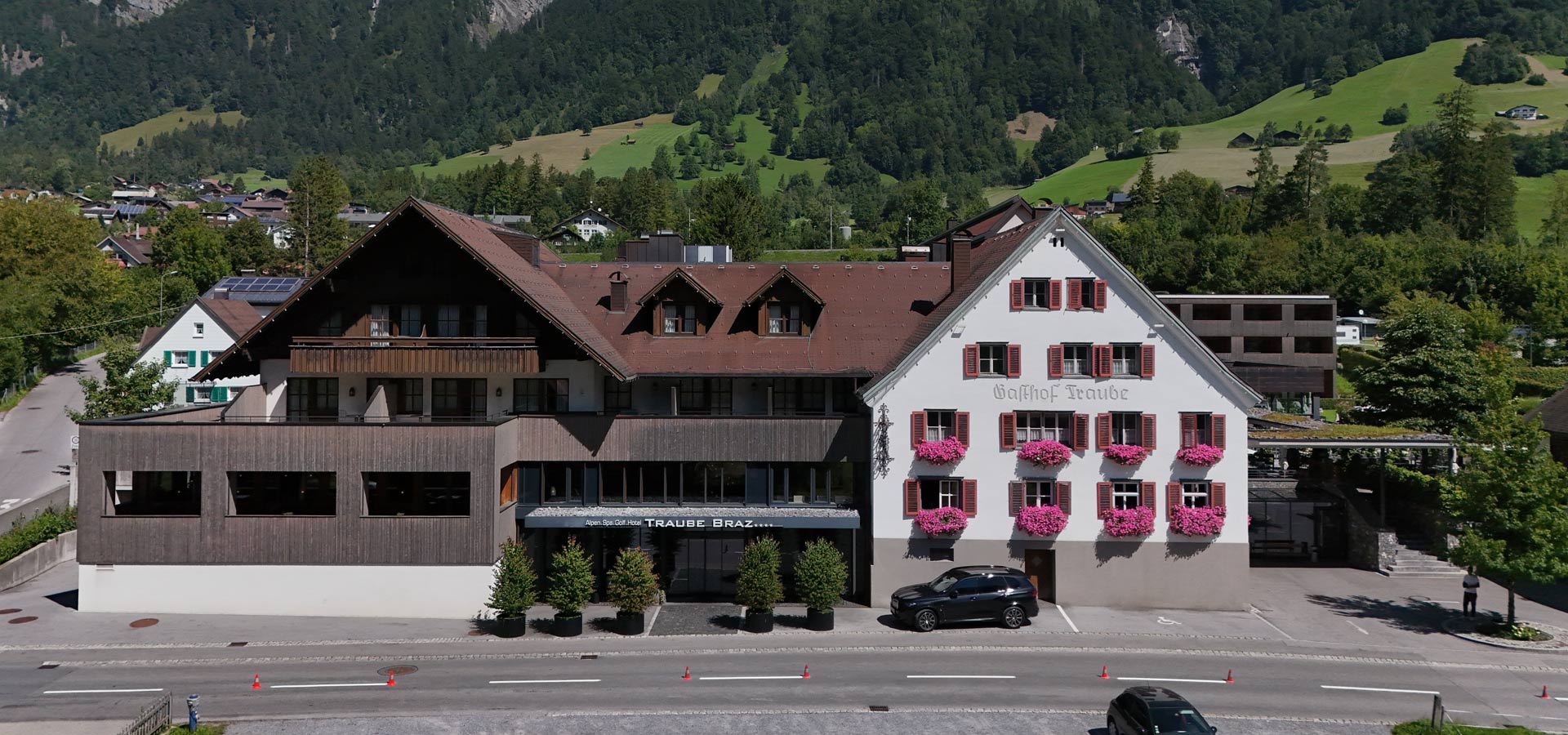Das Bild zeigt den Gasthof Traube, ein Gebäude mit zwei unterschiedlichen Fassaden. Die linke Seite ist aus Holz, während die rechte Wand weiß ist und mit roten Fensterläden sowie Blumenkästen geschmückt ist. Im Vordergrund sind Bäume und ein Parkplatz zu sehen.