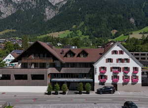 Das Bild zeigt ein Hotel im alpine Stil mit einem hellen, weißen Gebäude und einem dunklen Holzbereich. Es hat rote Fensterläden und Balkone mit Blumen. Im Hintergrund sind grüne Berge zu sehen, die die Idylle der Umgebung unterstreichen.