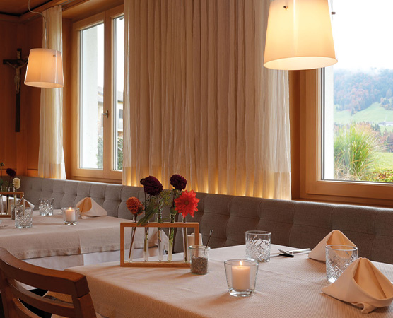 Ein gemütliches Restaurant mit einem gepolsterten Sitzbereich, in dem mehrere Tische mit Tischdecken gedeckt sind. Auf den Tischen stehen Kerzen und Blumenarrangements. Große Fenster sorgen für viel Tageslicht und bieten einen Blick auf die Natur draußen.
