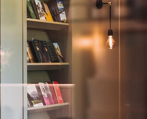 Ein Regal mit verschiedenen Prospekten steht im Hintergrund. Die Prospekte zeigen unterschiedliche Farben und Designs. Eine hängende Lampe beleuchtet den Bereich und schafft eine gemütliche Atmosphäre.