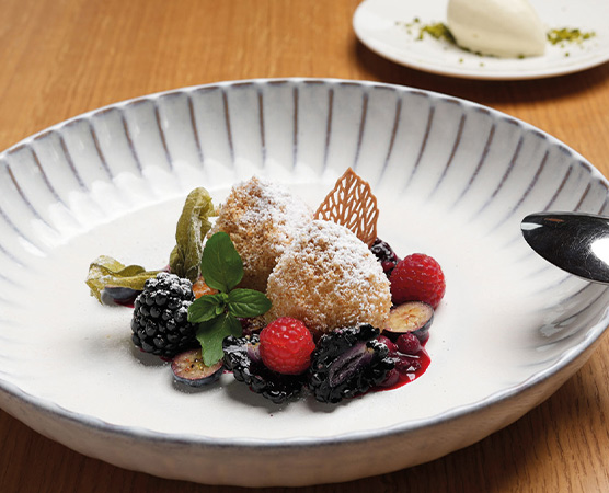 Ein tiefes, weißes Schüsselchen enthält zwei panierte Kugeln, bestreut mit Puderzucker, umgeben von einer Auswahl frischer Beeren wie Himbeeren, Heidelbeeren und Brombeeren. Frische grünes Blattwerk und eine dekorative Karamellnote ergänzen die Erscheinung des Desserts.