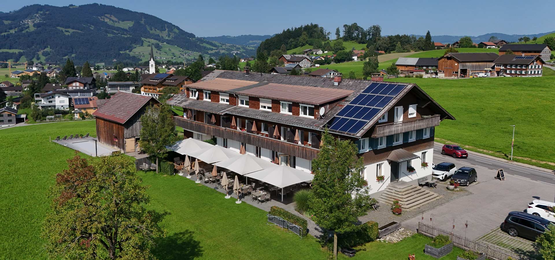Ein zweigeschossiges, traditionelles Holzgebäude mit einem modernen Dach voller Solarpaneele. Vor dem Haus befinden sich Terrassen mit Sonnenschirmen. Im Hintergrund erstrecken sich grüne Wiesen und sanfte Hügel. Die Umgebung strahlt eine ruhige, ländliche Atmosphäre aus.