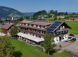 Das Bild zeigt ein Gebäude mit mehreren Stockwerken, das als Hotel erkennbar ist. Es ist von grünen Wiesen umgeben und hat eine Terrasse mit Sonnenschirmen. Auf dem Dach sind Solarpanels angebracht. Im Hintergrund sind sanfte Hügel und ein kleiner Ort zu sehen.