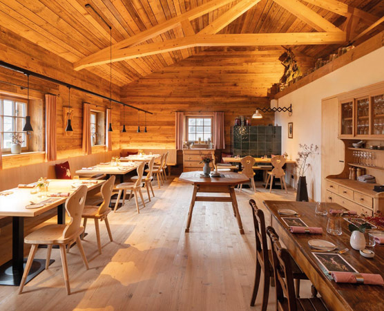 Das Bild zeigt ein gemütliches Restaurant mit holzvertäfelten Wänden und einem offenen Dachs. Mehrere Esstische sind im Raum verteilt, umgeben von Stühlen. Eine entspannte Atmosphäre wird durch die warmen Farben und natürliche Materialien vermittelt. Ein Fenster lässt Tageslicht herein.