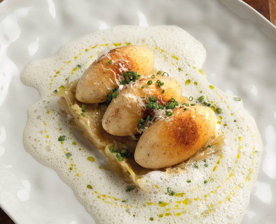 Ein kunstvoll angerichteter Teller mit drei gebräunten, ovalen Klößen, die auf einer Basis aus zartem Gemüse liegen. Umrahmt von einer schaumigen Sauce, sind die Klöße mit feinen grünen Kräutern und einer Prise Gewürz verziert, was dem Gericht einen eleganten Look verleiht.